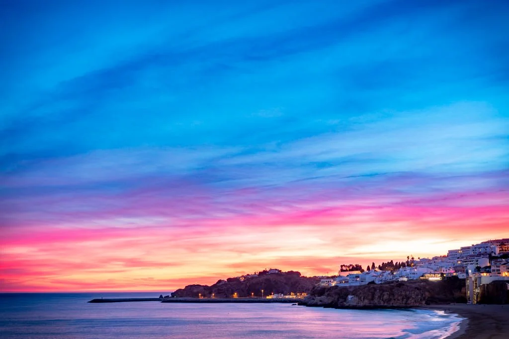 Colorful sunset over the ocean with a hillside town on the right, white buildings, and a pier stretching into the water.