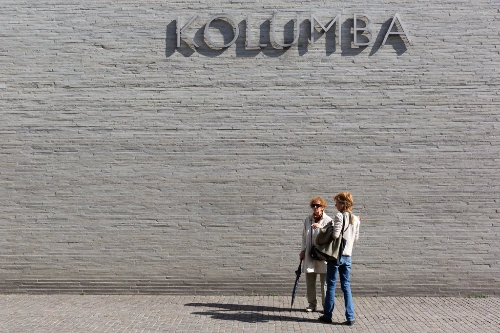 Two women talking and standing against a large white brick wall with the word 'KOLUMBA' in large letters.