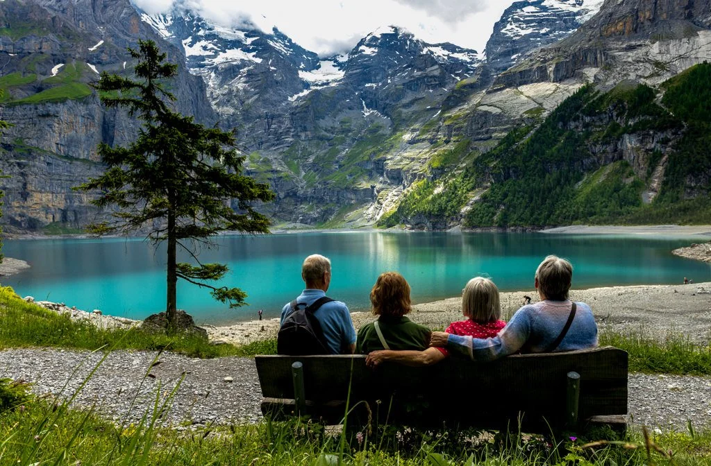 Four people sitting on a bench by a turquoise lake in a mountainous landscape with greenery and snow-capped peaks.