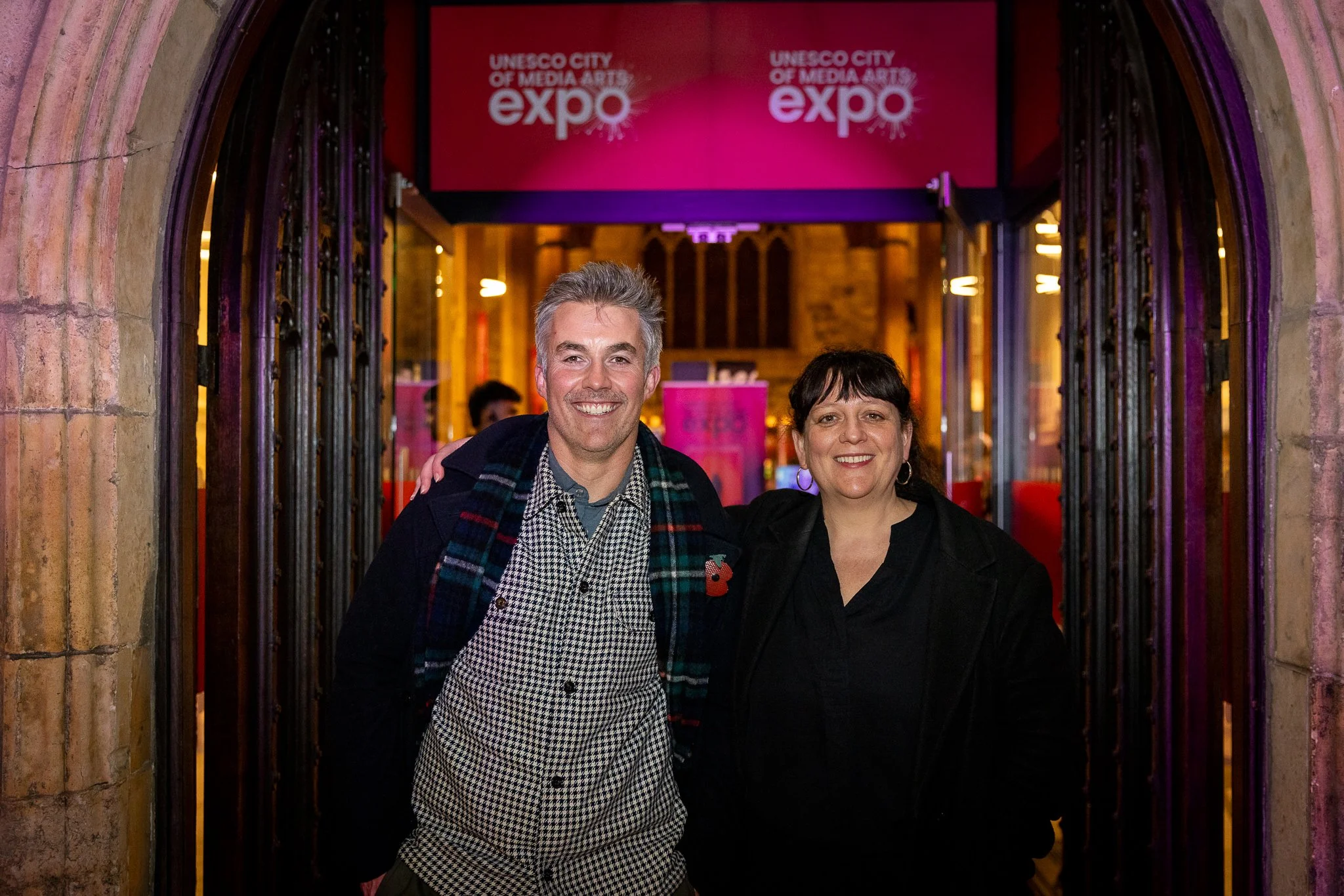 Two smiling people standing in front of the entrance to the UNESCO City of Media Arts expo, with a pink sign overhead and arched doorway framing them.