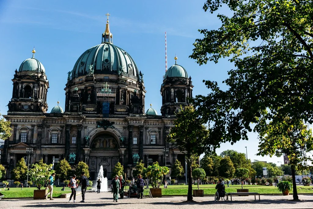 The Berlin Cathedral with a large green dome, surrounded by trees and people walking in the park.