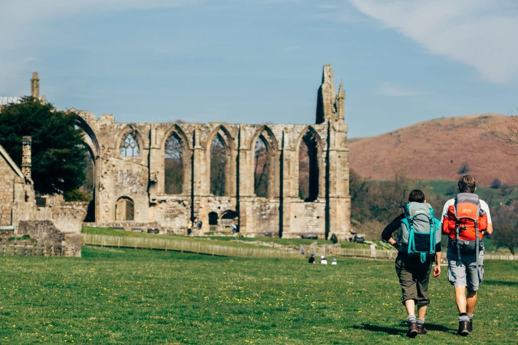 Two hikers with backpacks walking toward the ruins of an old stone church or abbey, with hills in the background.
