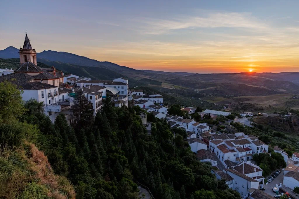 Sunset over a hillside village with white buildings and terracotta roofs, surrounded by greenery and mountains in the distance.