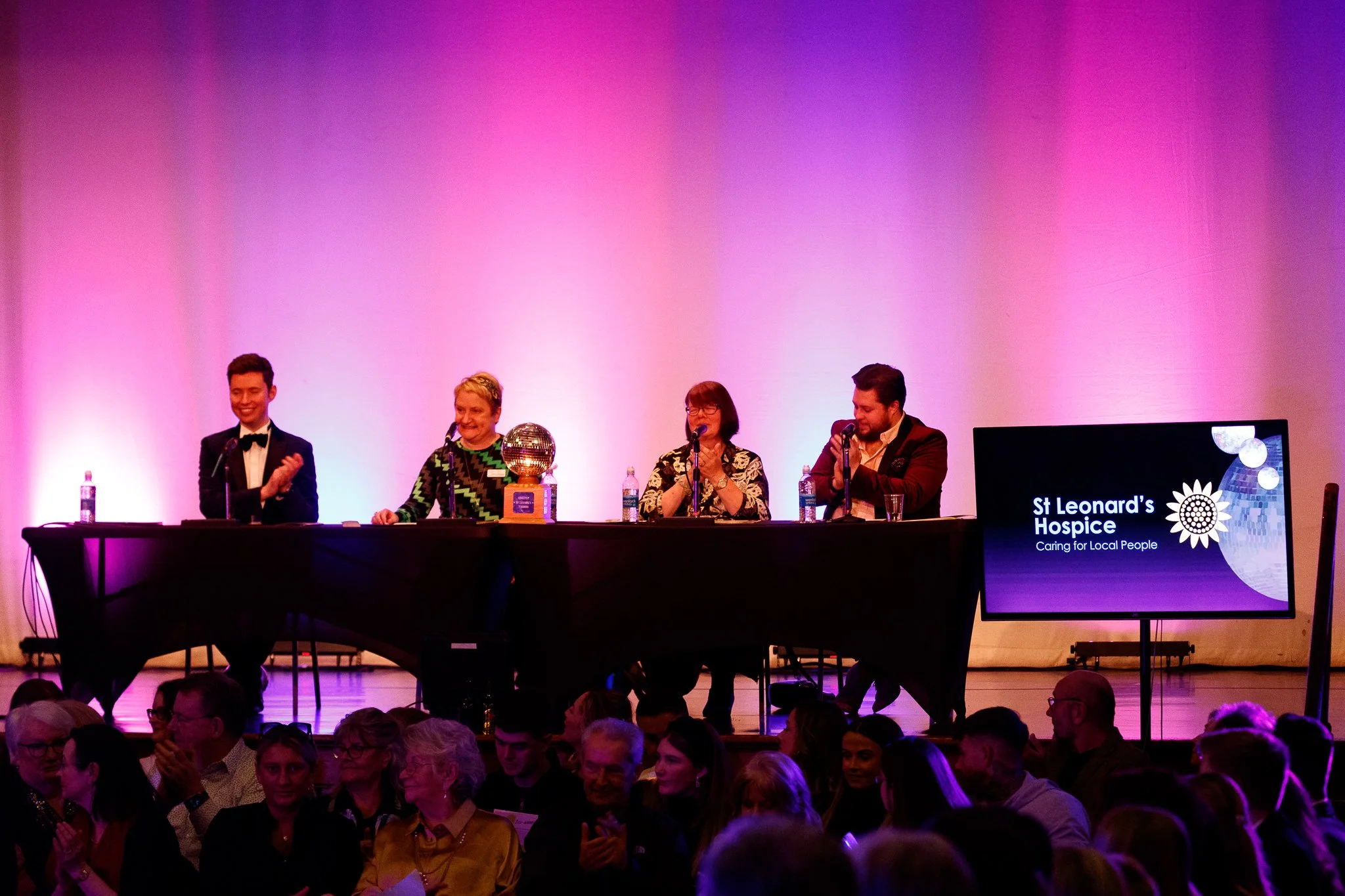 A panel of five people on a stage at an event with a pink and purple background. The panel includes three women and two men, with water bottles on the table in front of them. There is a large screen displaying the logo of St Leonard's Hospice and the