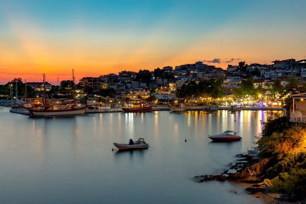 Sunset over a marina with boats and a hillside town illuminated by streetlights