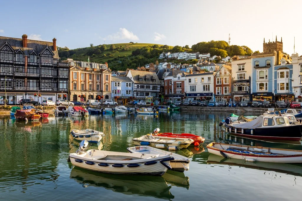 A harbor scene with several boats docked on calm water, a row of colorful buildings along the waterfront, and a hill with houses in the background.