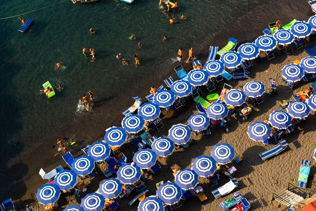 Beach scene with numerous blue and white striped umbrellas, lounge chairs, and people swimming and relaxing in the water.