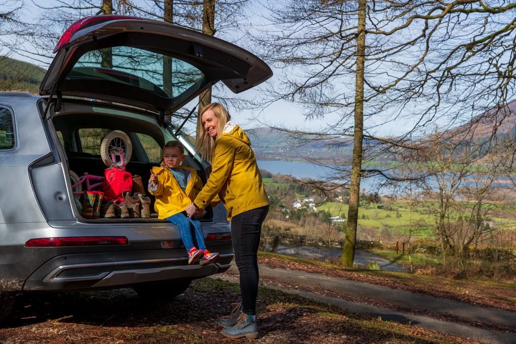 A woman helping a young girl get into the back of a car during a packed outdoor trip, with a scenic lake and mountains in the background.