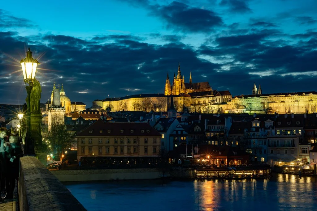 Night view of Prague with illuminated Prague Castle and Charles Bridge over the river, cloudy sky, and streetlights reflecting on the water.