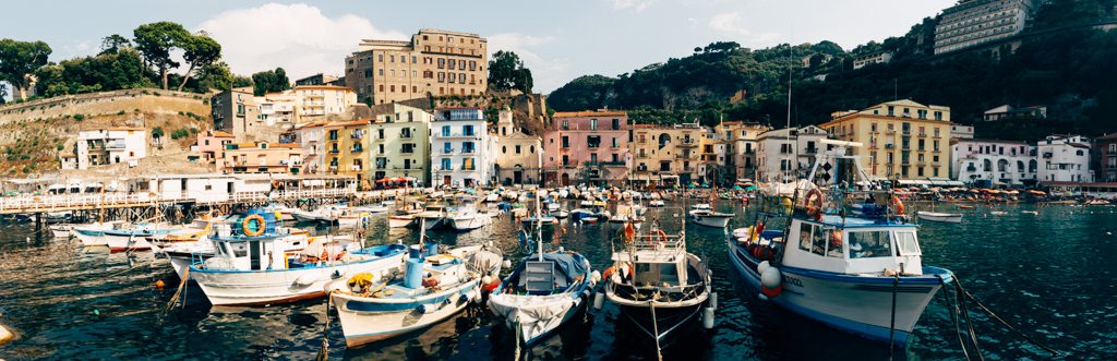 Colorful buildings along a harbor with several boats docked in the water, hillside with trees in the background.