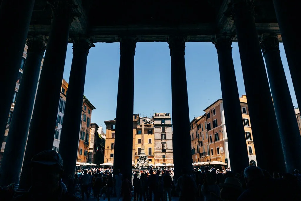 Looking up at the Colosseum in Rome, Italy, with tall columns framing the scene and a crowd of tourists in front of historic buildings.