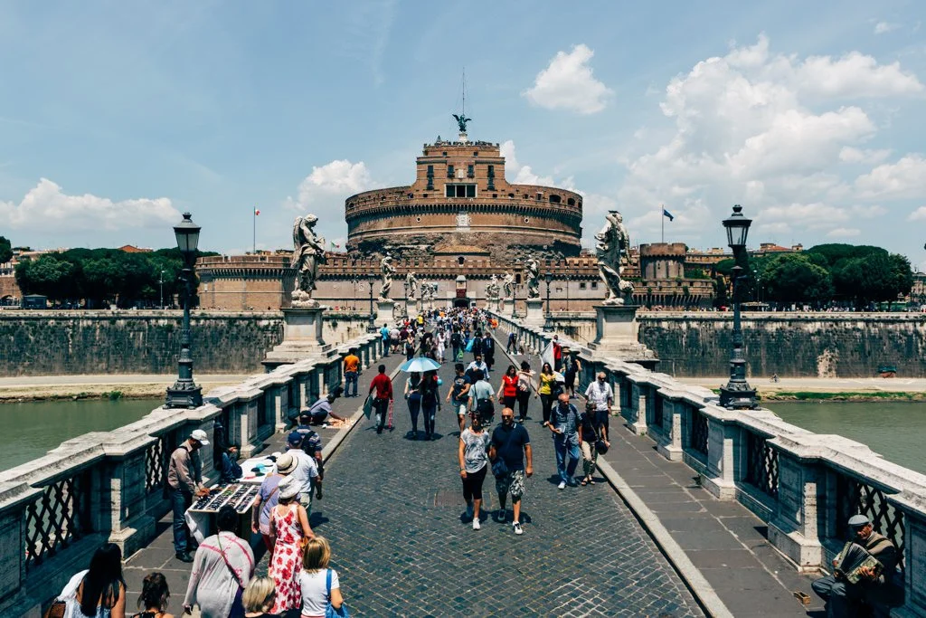 People walking across the Tiber River on the Ponte Sant'Angelo bridge towards Castel Sant'Angelo in Rome, Italy, with statues and historic architecture visible.