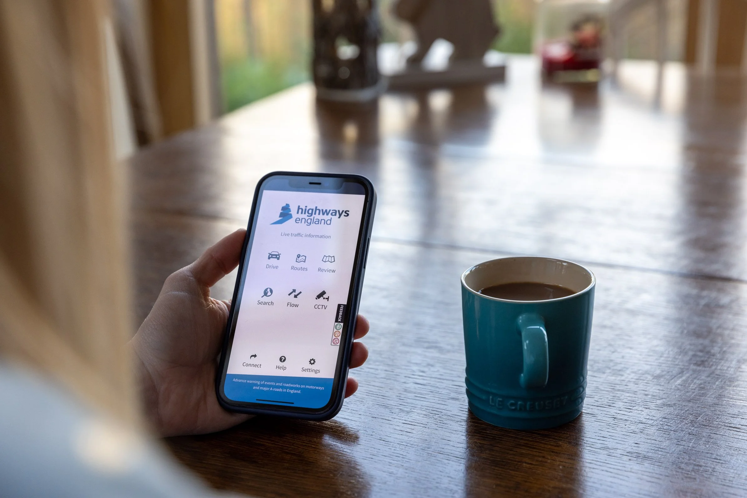 Person holding a smartphone with highways england app open, sitting at a wooden table with a blue mug filled with coffee or tea. In the background, blurred natural scenery seen through a window.