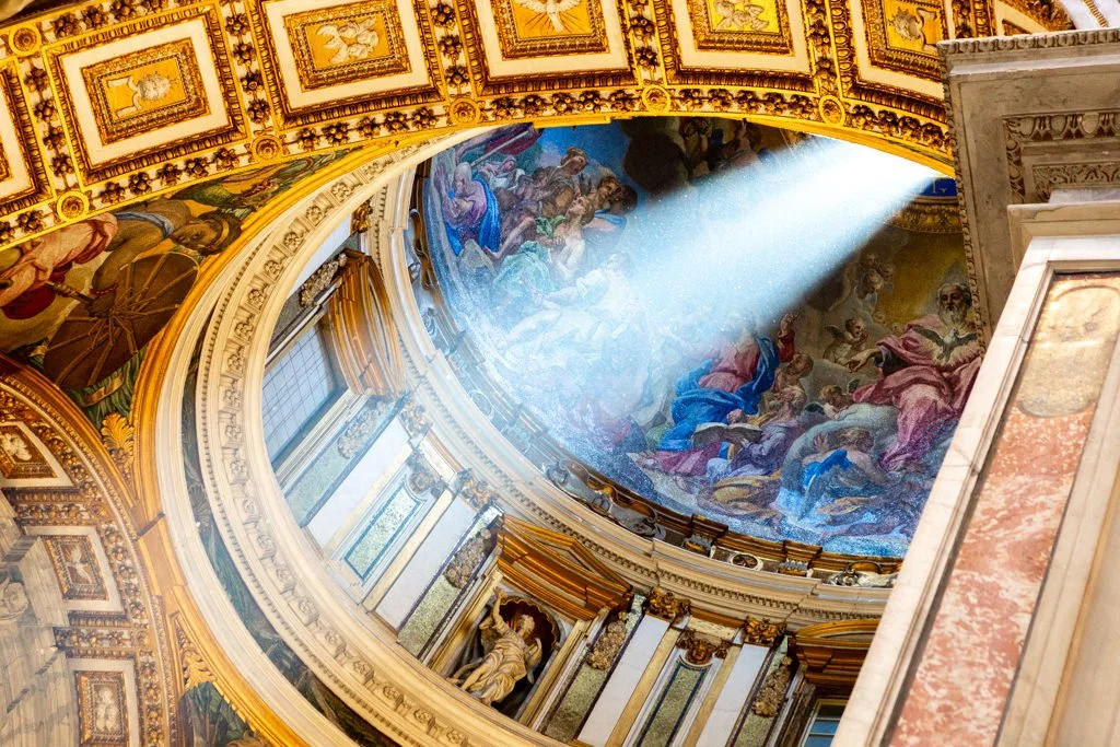 Interior view of a grand, ornate domed ceiling with vibrant religious paintings and a beam of sunlight shining through.