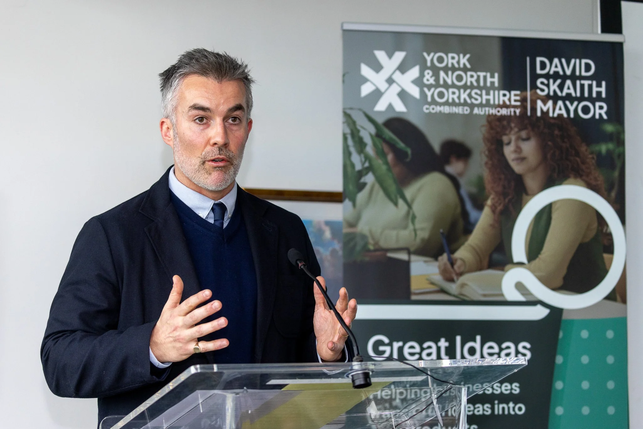 A man with gray hair and a beard wearing a dark suit and tie speaking at a podium during an event, with a York & North Yorkshire Combined Authority banner in the background.