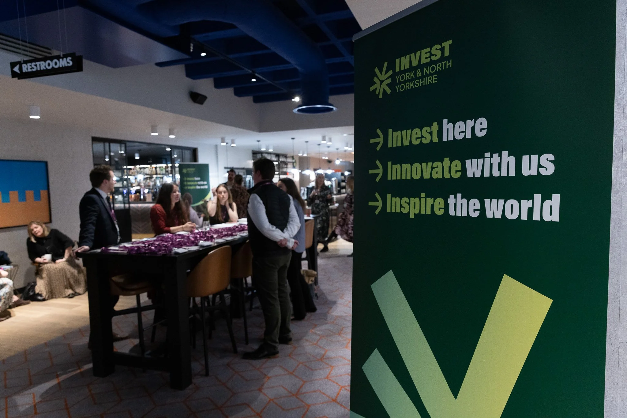 A group of people gathered around a table with purple decorations in a modern indoor setting, with signs promoting investment, innovation, and inspiration from Invest York & North Yorkshire.