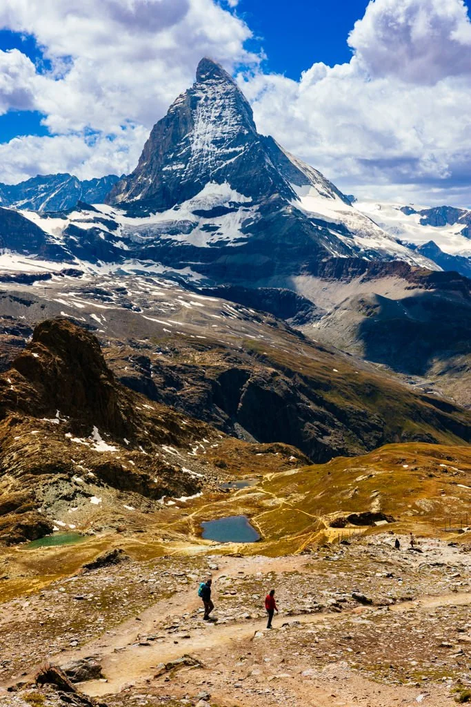 Two hikers walk along a rocky trail in a mountainous landscape with a snow-capped peak towering in the background. Small lakes are visible in the foreground amidst patches of snow and grass.