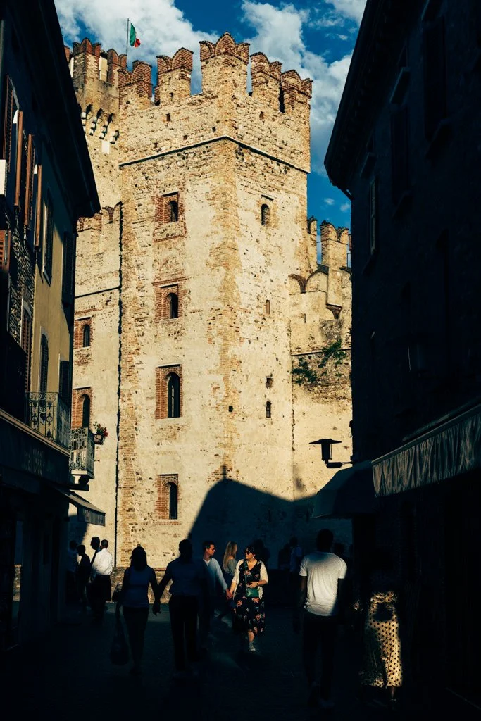 People walking in a narrow street with a large medieval stone castle with battlements and an Italian flag in the background.
