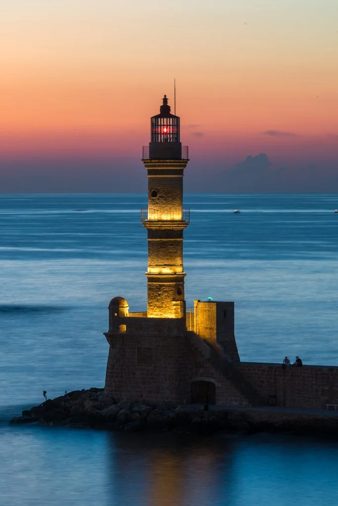 A lighthouse illuminated at sunset by the sea with a colorful sky in the background.