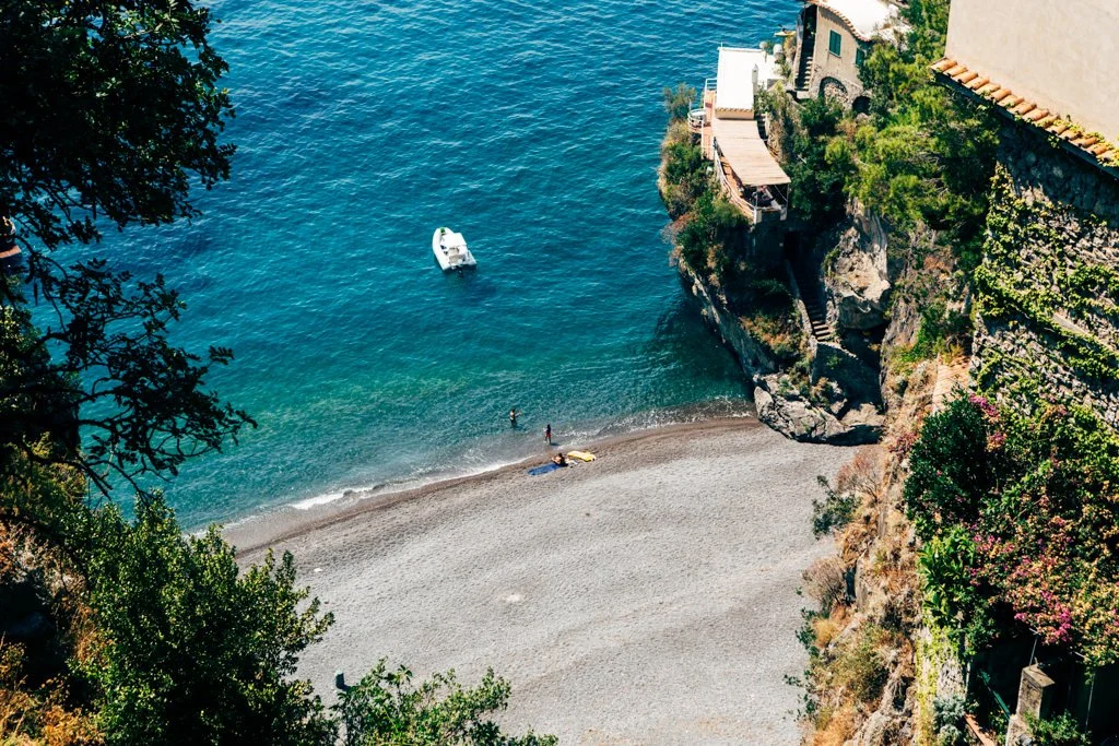 A small pebble beach with two people and a boat floating on the water, surrounded by greenery and buildings on a cliffside.
