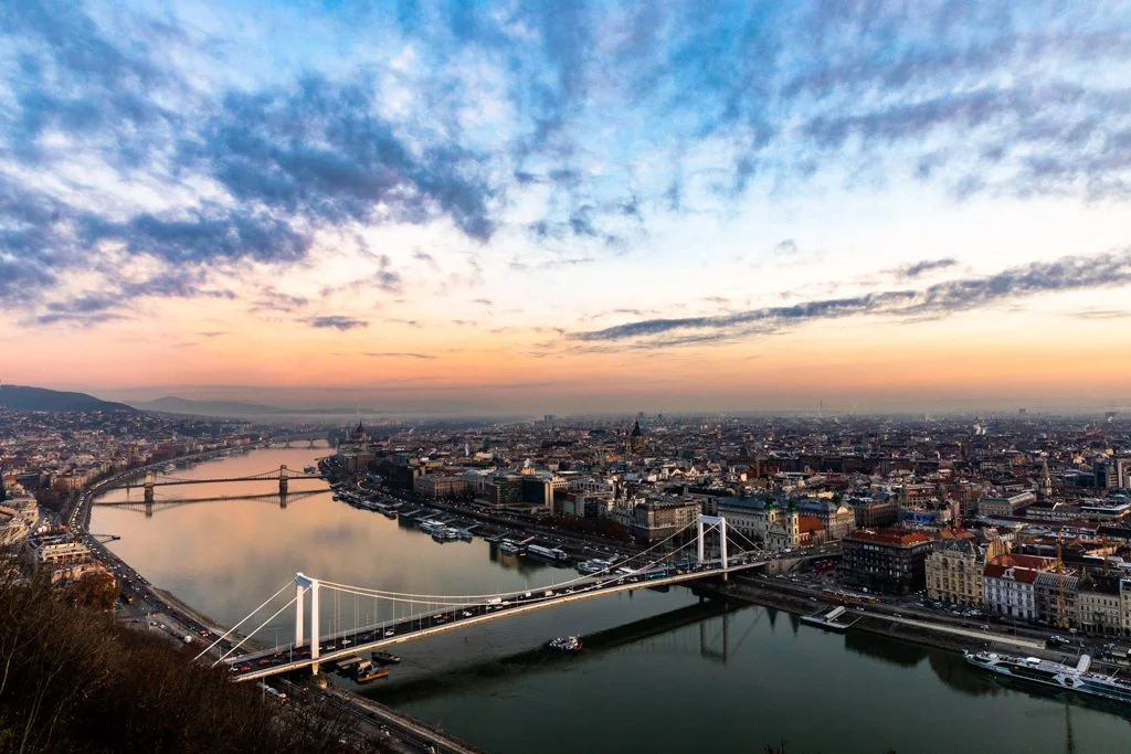 A city skyline at sunset with a river running through the city. There is a bridge crossing the river in the foreground and several buildings and hills in the distance.