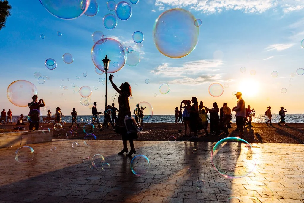 People on a beach at sunset with bubbles, some people taking photos, and a woman playing with bubbles near a lamp post.