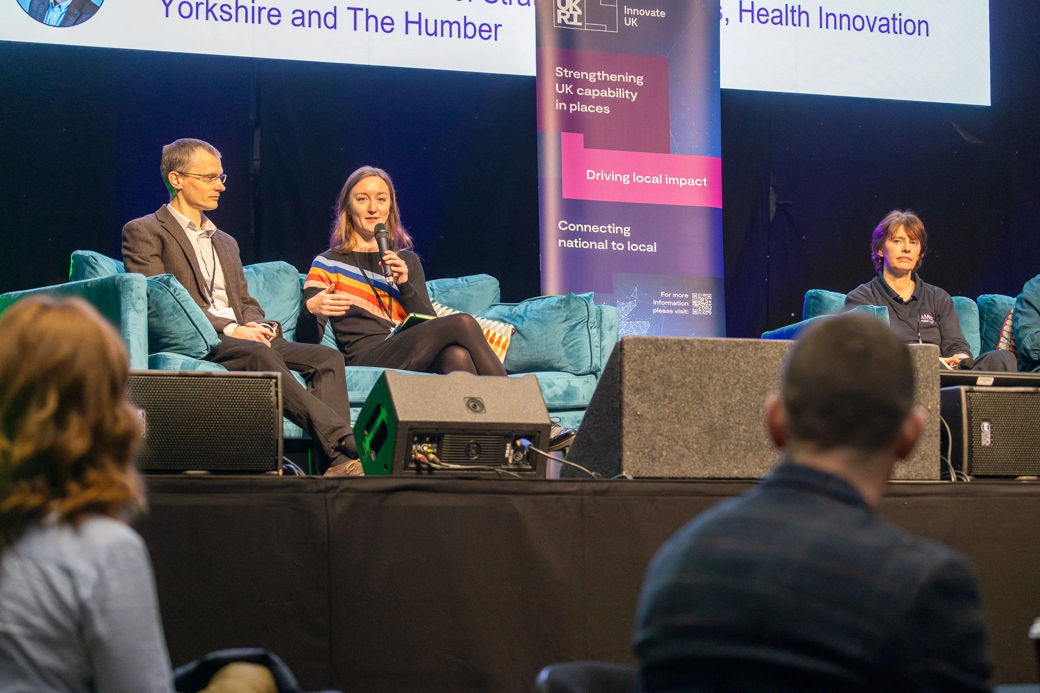 Three panelists seated on a stage during a conference, one woman speaking into a microphone, with a large presentation screen behind them displaying text about UK health innovation and regional development.