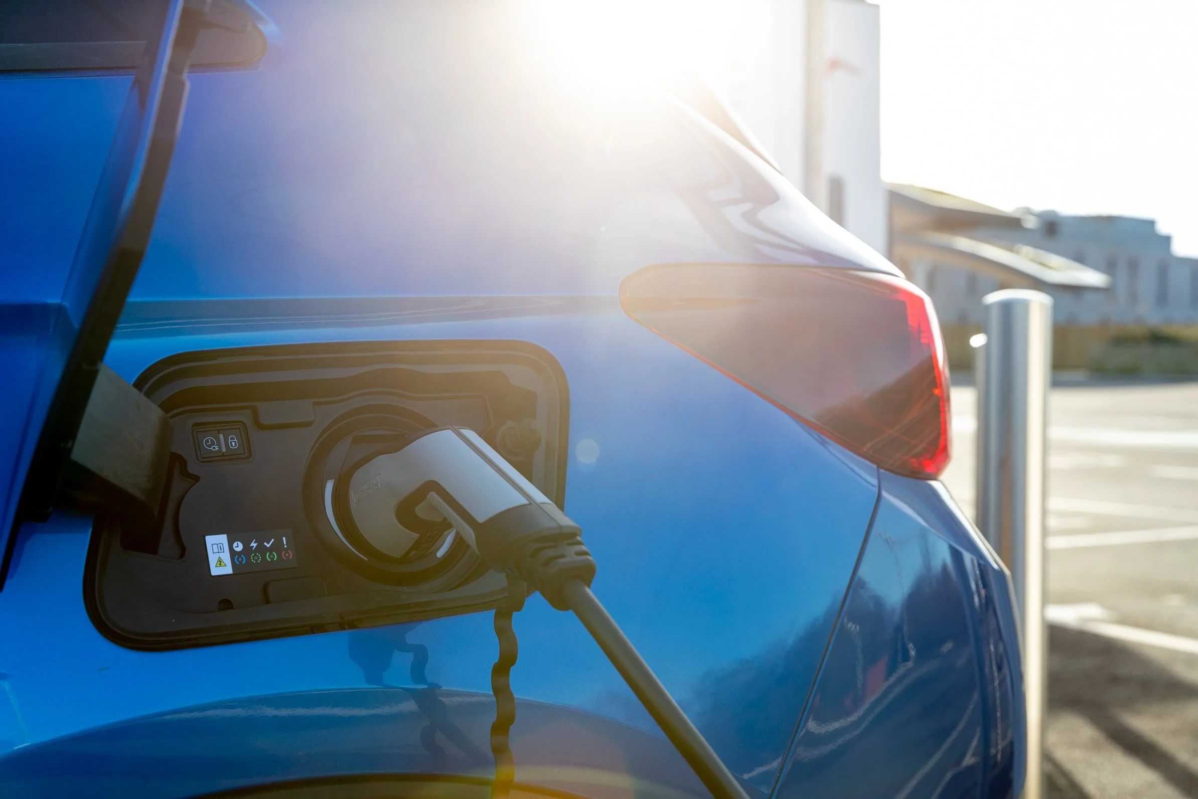 Close-up of electric vehicle charging port with charging cable plugged in, sunlight reflecting off vehicle's surface, and an urban background with buildings.