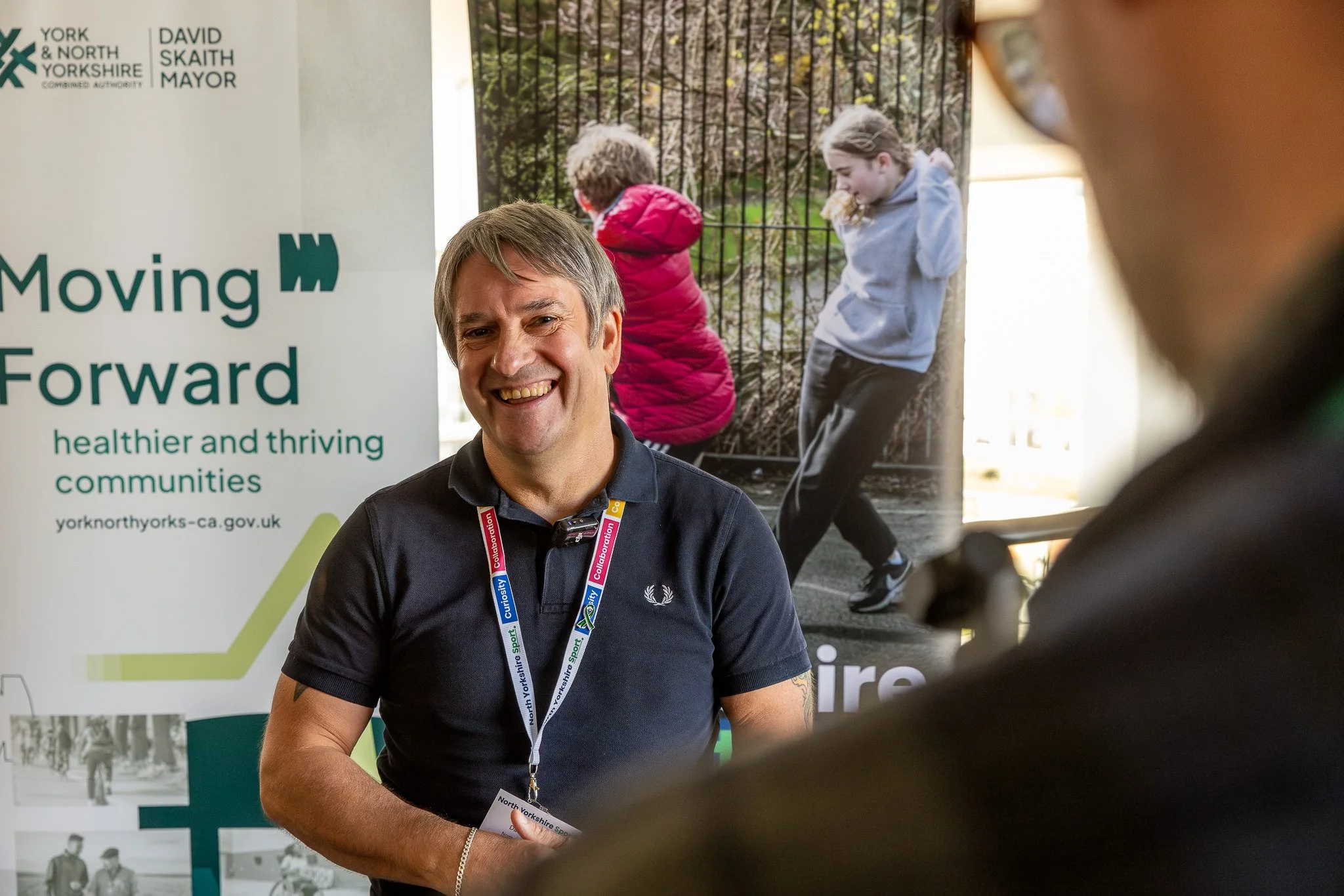 Smiling man with gray hair, wearing a black polo shirt and lanyard, standing in front of a poster that reads 'Moving Forward' with images of community scenes and two children.
