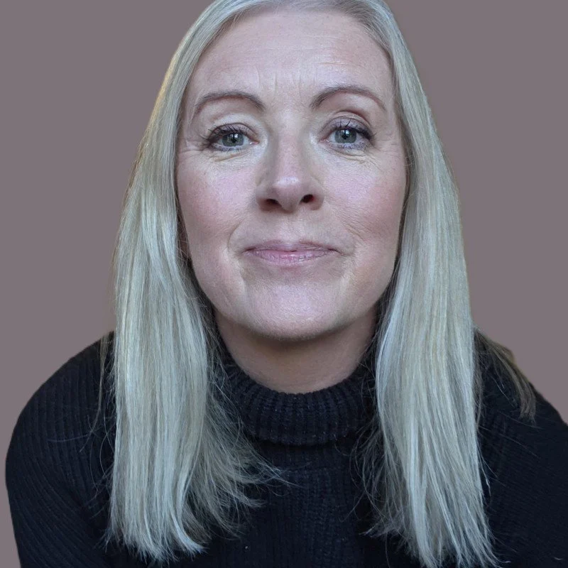 Close-up of a middle-aged woman with long blonde hair, blue eyes, wearing a black turtleneck sweater, smiling softly against a plain background.