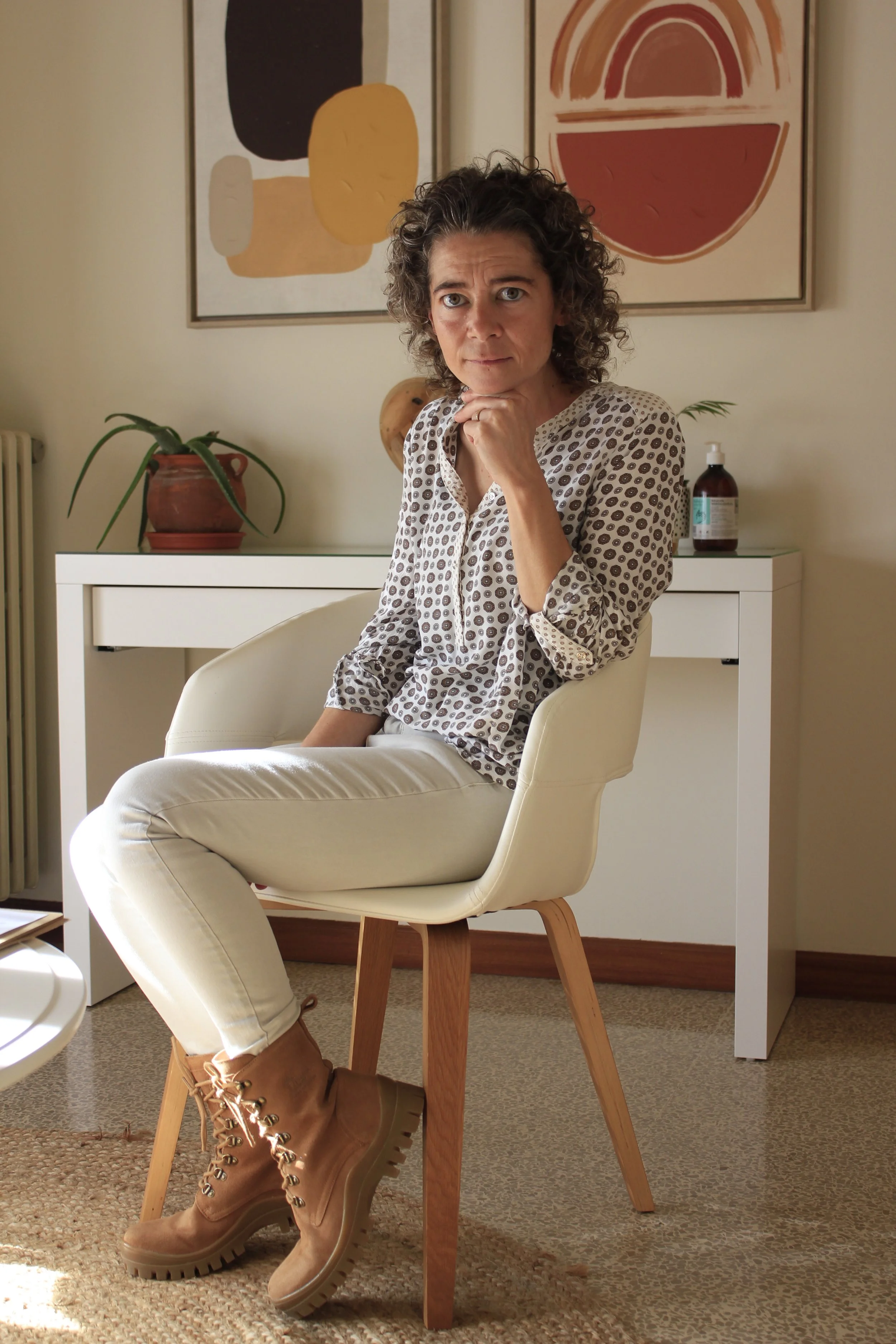 Psicoterapeuta sentada en una silla blanca con un gesto pensativo, en un espacio interior decorado con arte abstracto, plantas y artículos de madera.