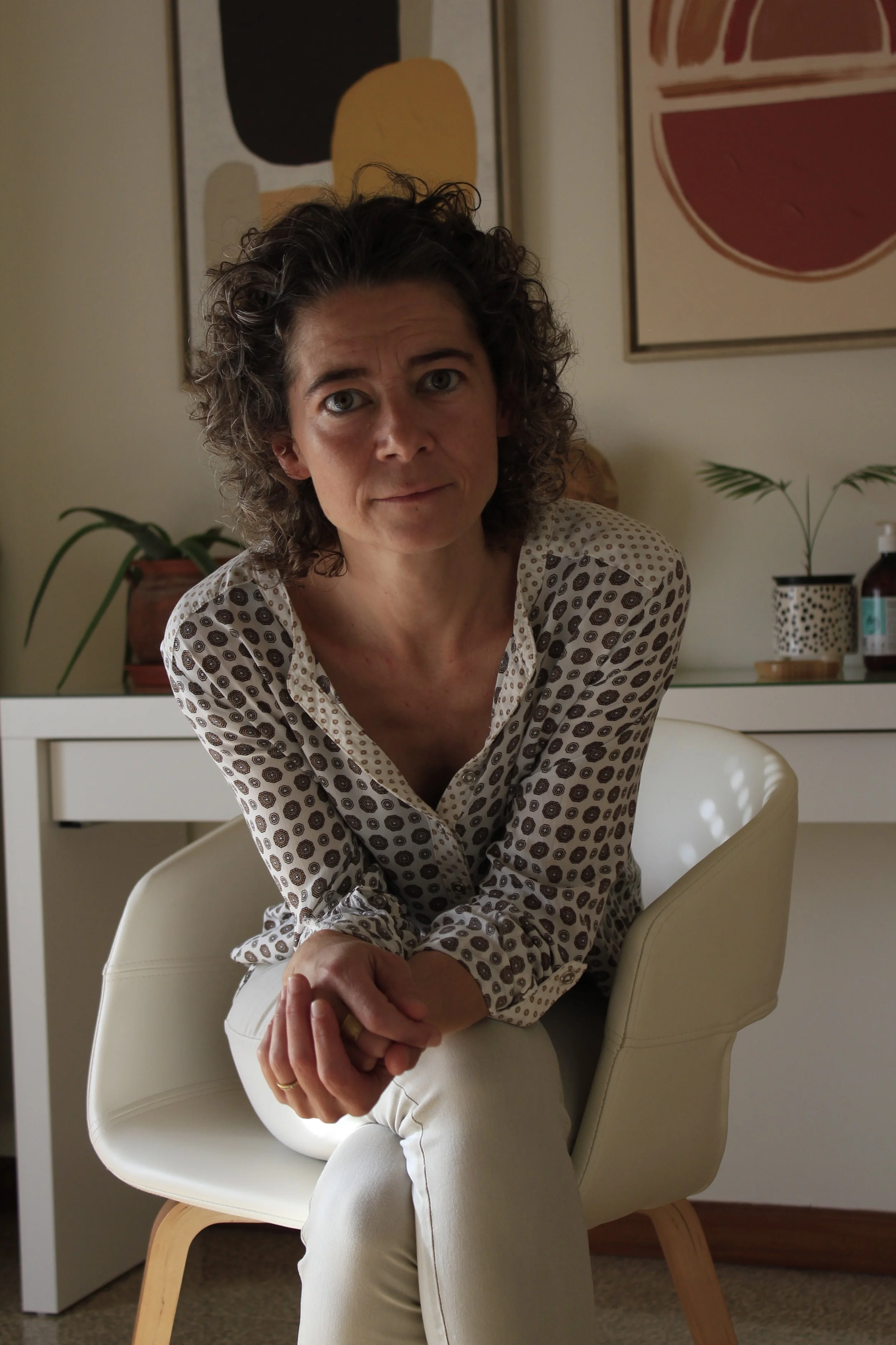 psicoterapeuta con cabello rizado sentado en una silla blanca en el interior de la consulta decorada con arte moderno y plantas.