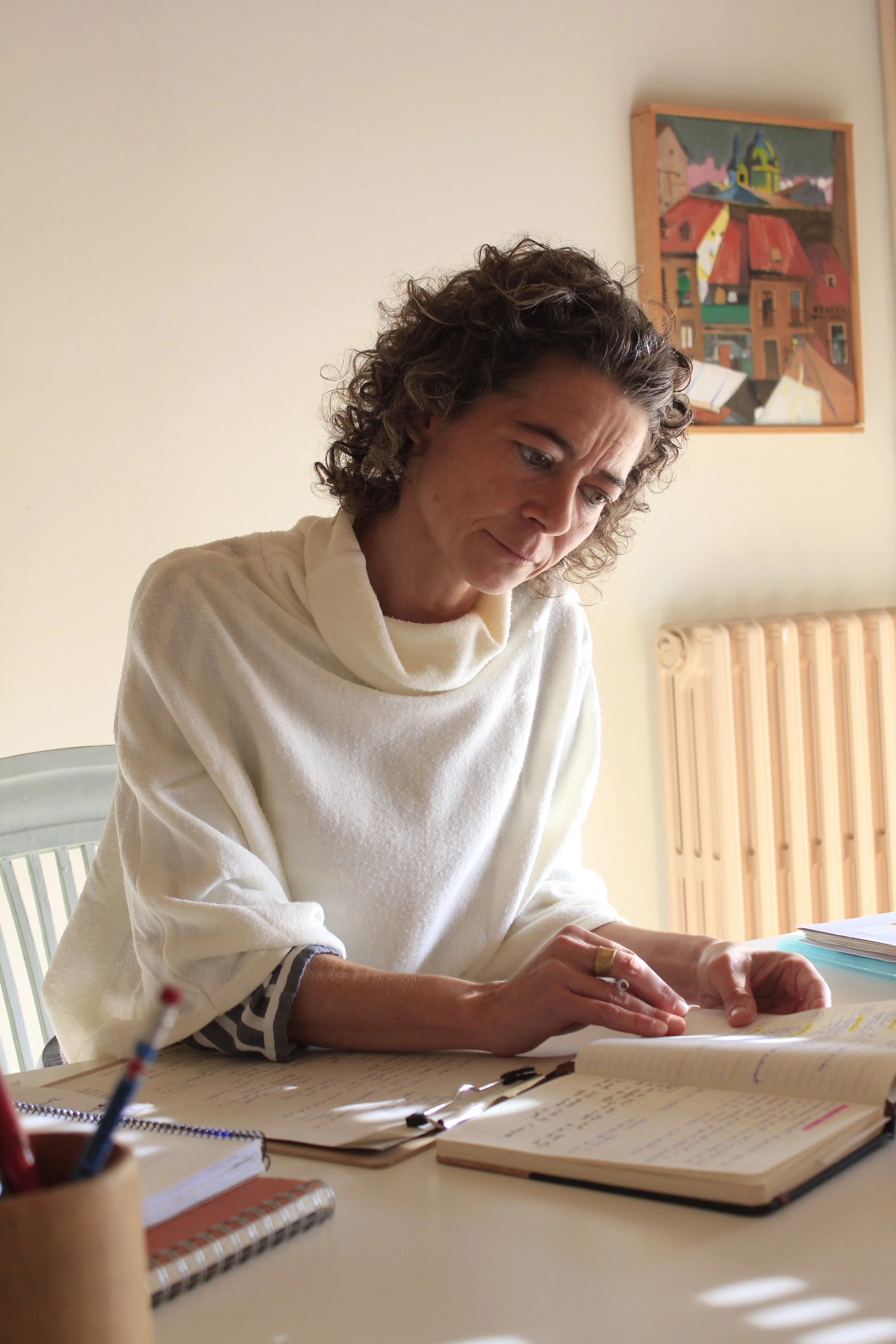 Mujer con cabello rizado leyendo notas en una mesa, en un ambiente interior con pintura colorida en la pared detrás.