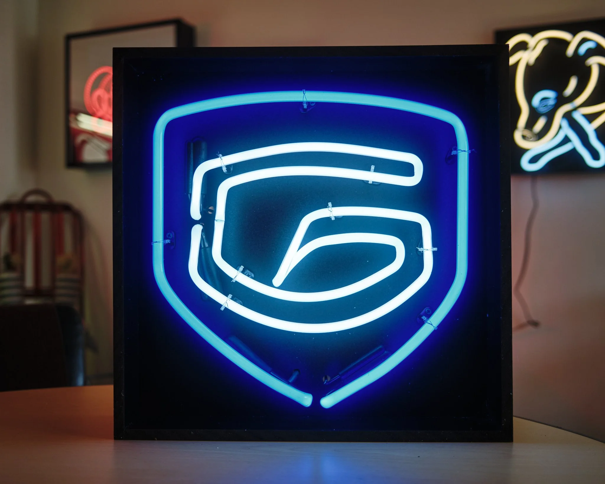 Glazton Auto Glass's logo (a stylized G shaped like a car windshield within a blue shield) created in neon tubing.  Featuring 12mm royal blue for the shield and 10mm cool white for the G, this logo measures 16.5" square and can be displayed on a book