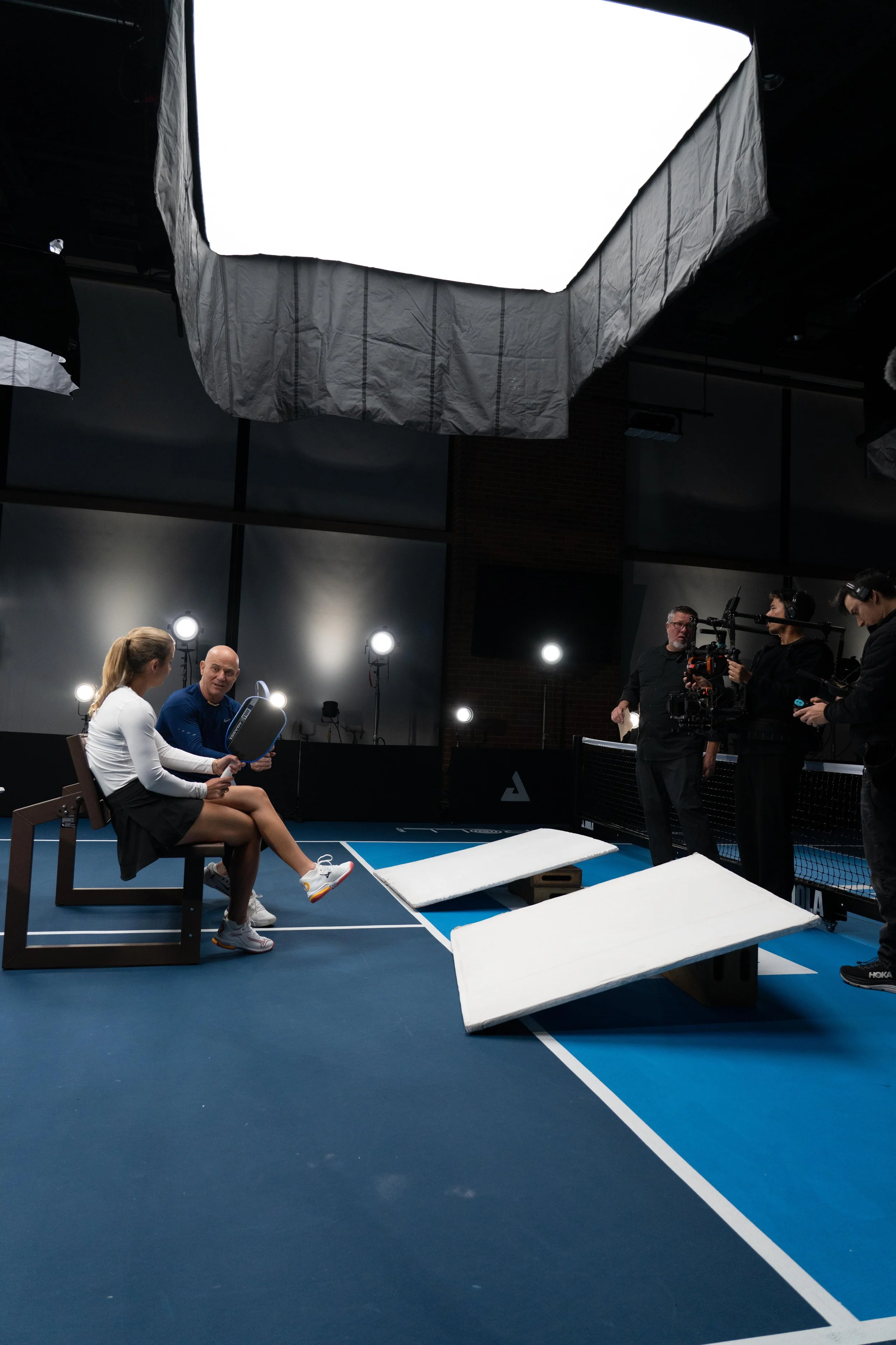 Indoor tennis court with a large studio light on a stand, two women sitting on chairs having a conversation, and a man operating a camera, with photography equipment and foam mats on the floor.