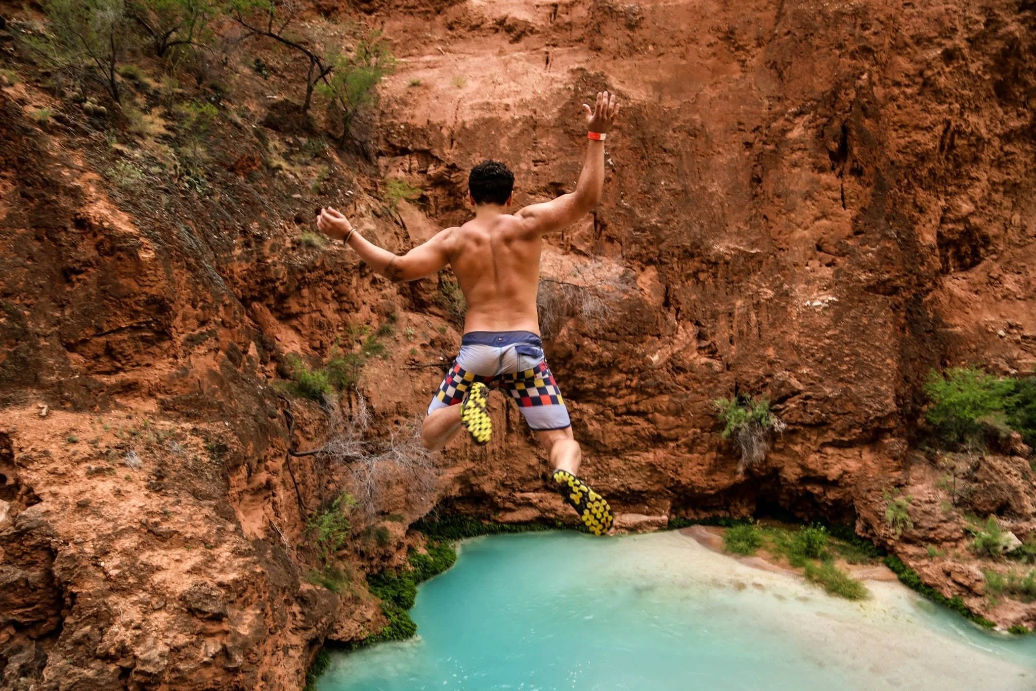 A shirtless man wearing colorful shorts and yellow climbing shoes, equipped with a climbing harness and safety gear, is mid-air jumping over a canyon with reddish rock walls and a body of turquoise water below.
