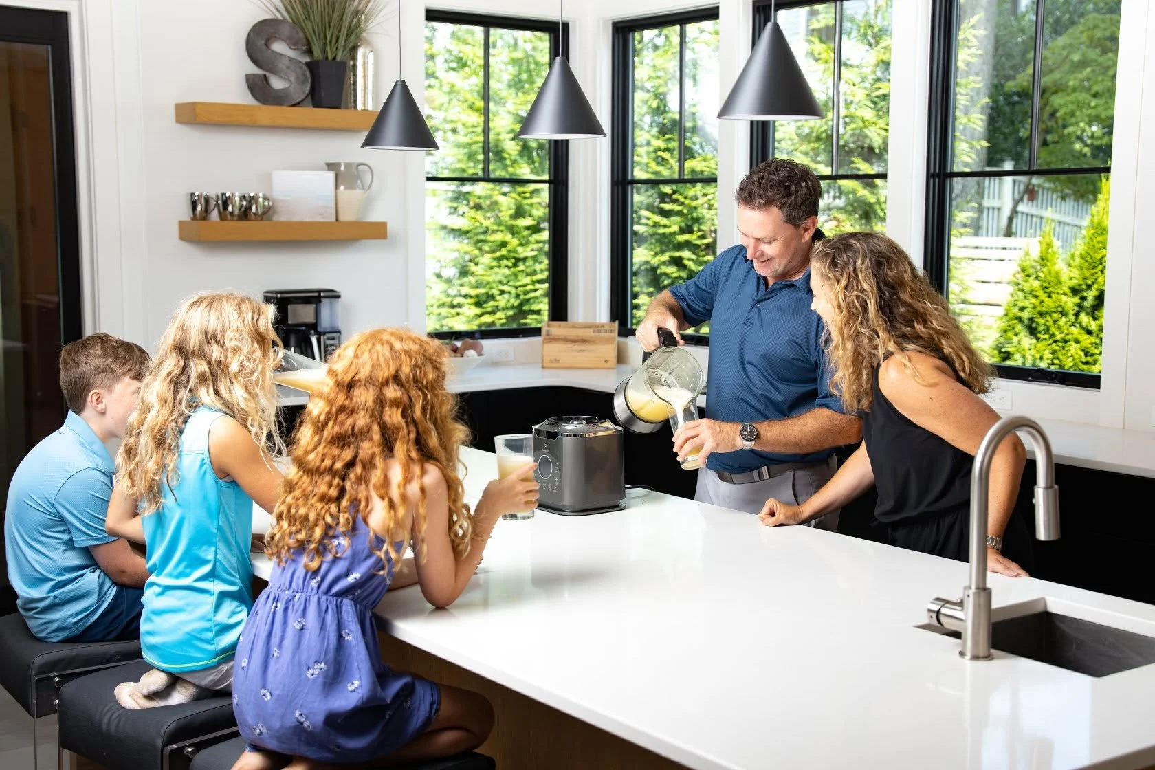 People in a kitchen with large windows, children sitting on barstools, adults pouring drinks, and a blender on the counter.