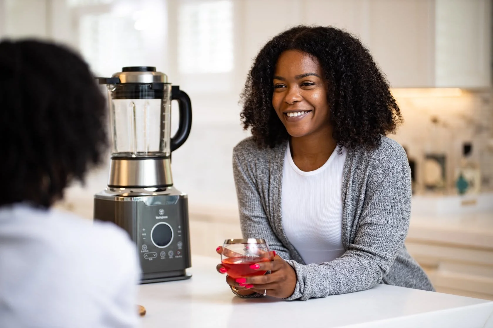 A woman with curly hair smiling and holding a glass of red wine while sitting at a kitchen counter, with a blender nearby.