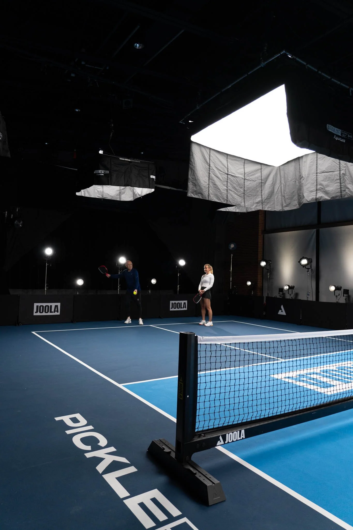 Indoor pickleball court with two players holding paddles and a ball, surrounded by professional lighting equipment and a net, branded with JOOLA branding.