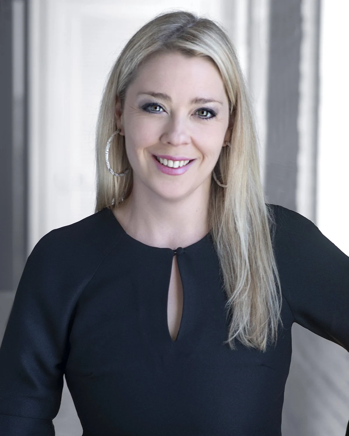 A woman with blonde hair, wearing hoop earrings and a black top with a keyhole neckline, smiling in front of a modern background.