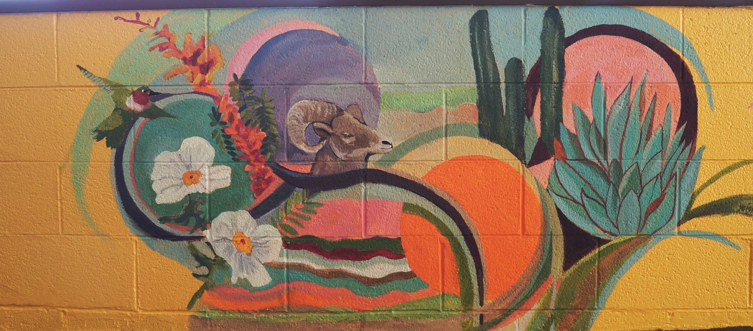 Colorful mural painting featuring a hummingbird, a ram's head, flowers, leaves, and abstract shapes on a yellow wall.