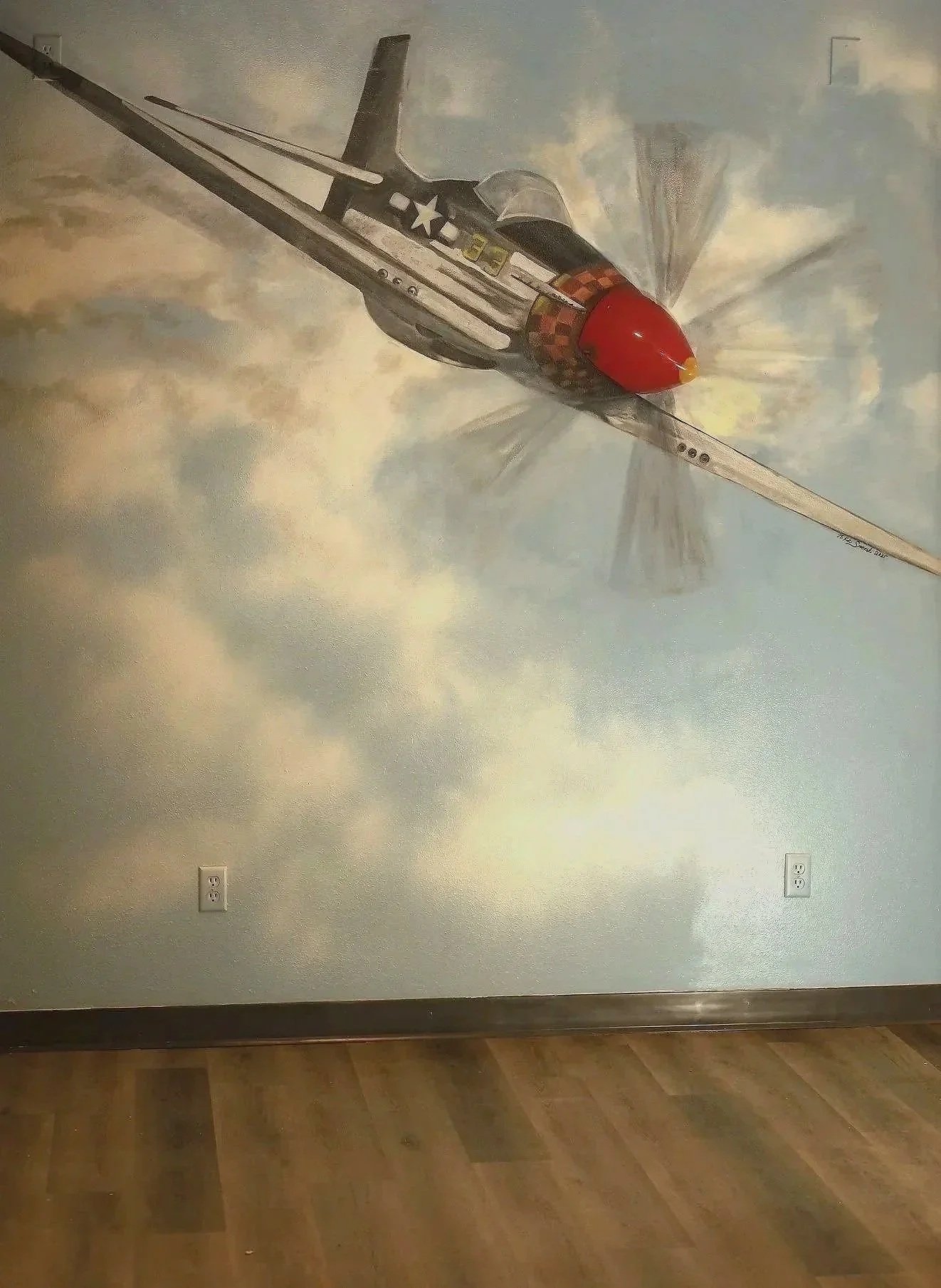 Wall mural painting of a fighter jet in mid-flight with clouds in the background, located on a wall above electrical outlets and a wooden floor.