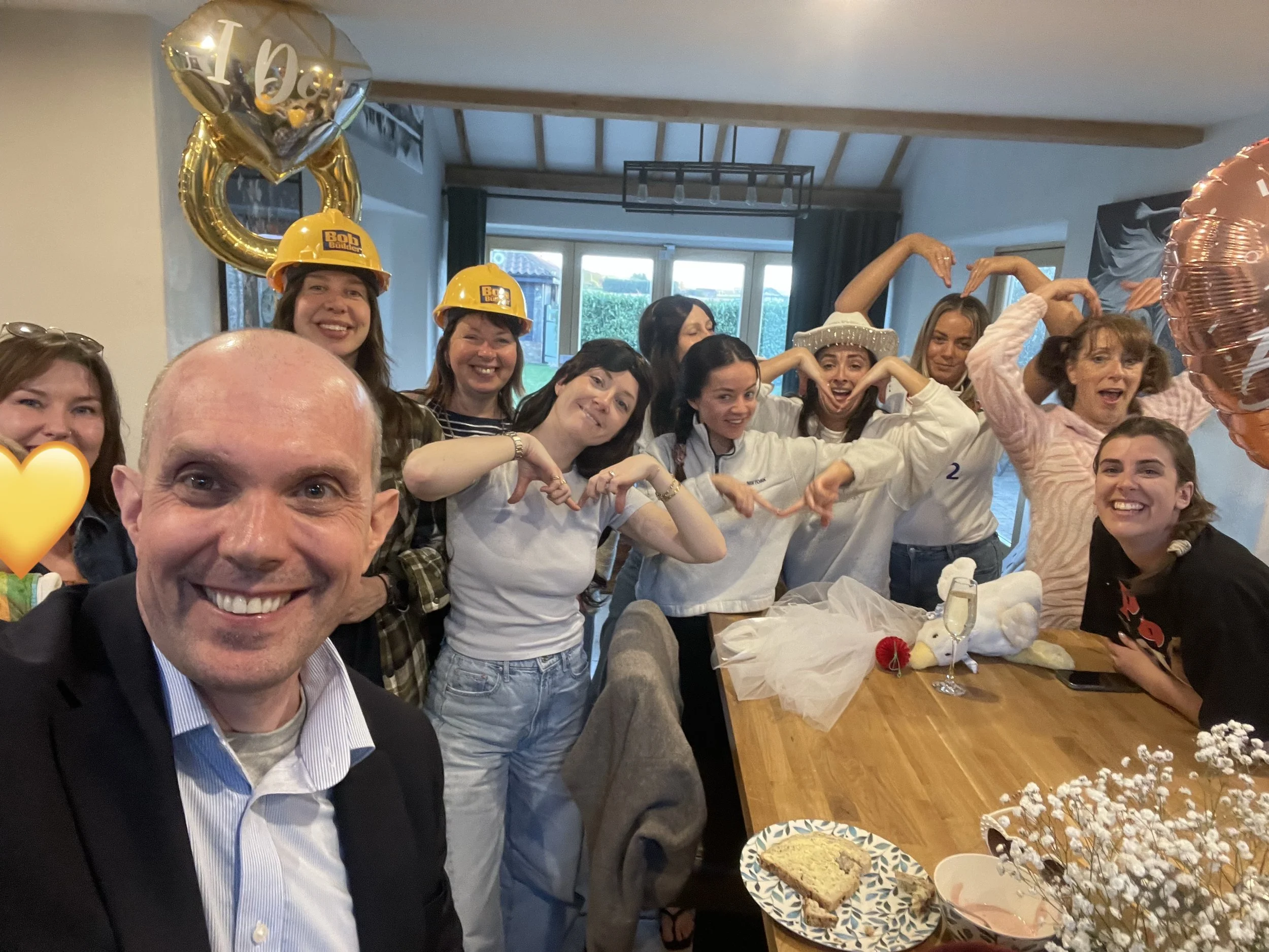 Group of women celebrating a private party, with Norwich magician David Richards in the foregroun, taking a selfie. They are all smiling and wearing fancy dress, with many making heart shapes with their arms, with balloons and a man taking a selfie.