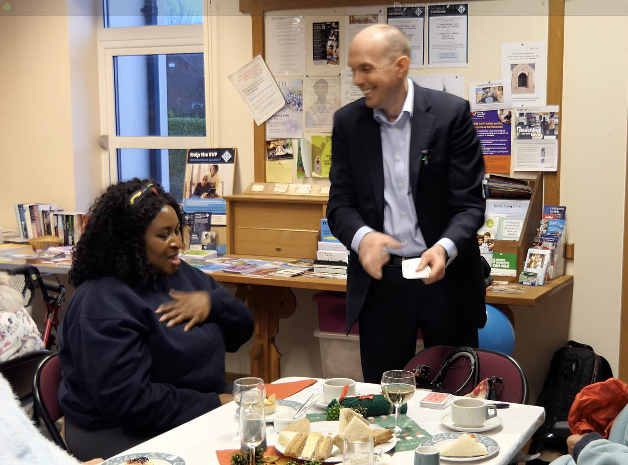 A woman sitting at a table with food and drinks, looking at a man standing and smiling in a room decorated with books, posters, and brochures.