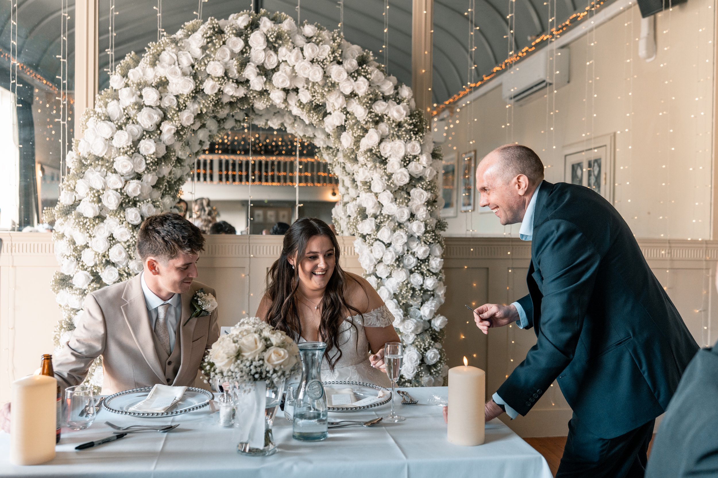 David Richards is performing for a  bride and groom sitting at a decorated table at a wedding reception. The bride is  laughing, and the groom is smiling.