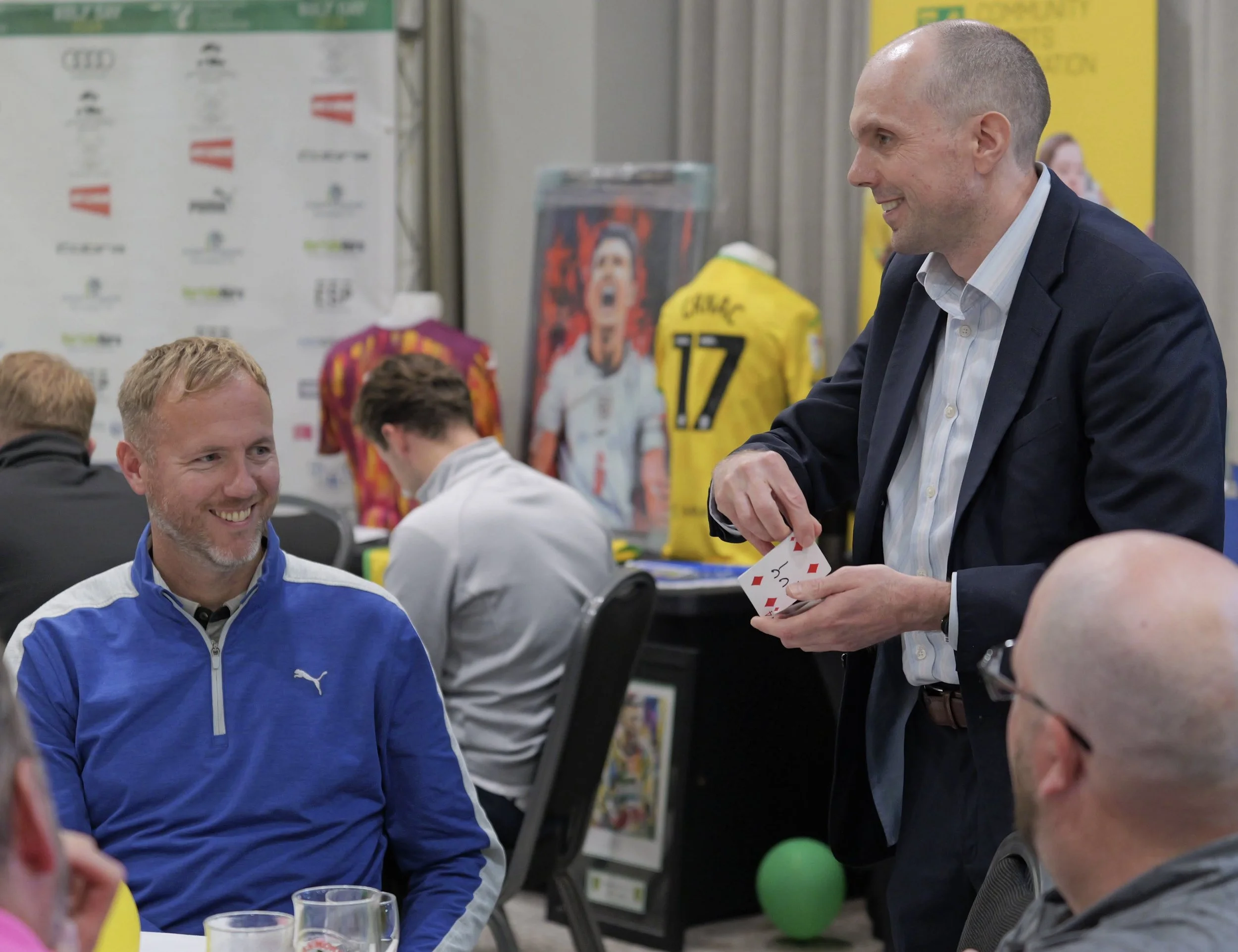 Norwich magician David Richards performing a card trick for a group of people sitting at a table during an indoor event.