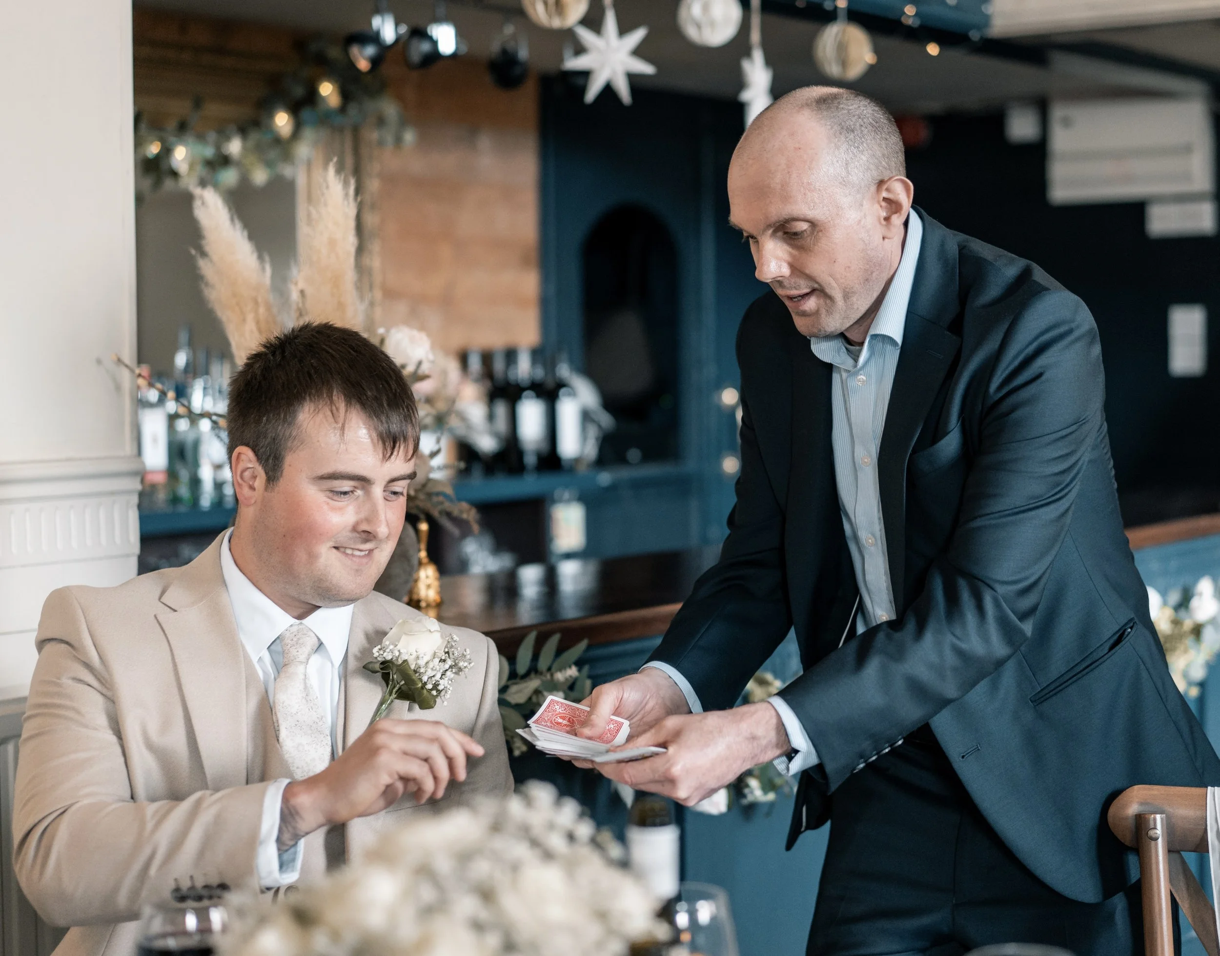 David Richards performs magic to a seated guest at a wedding, asking him to choose a playing card.
