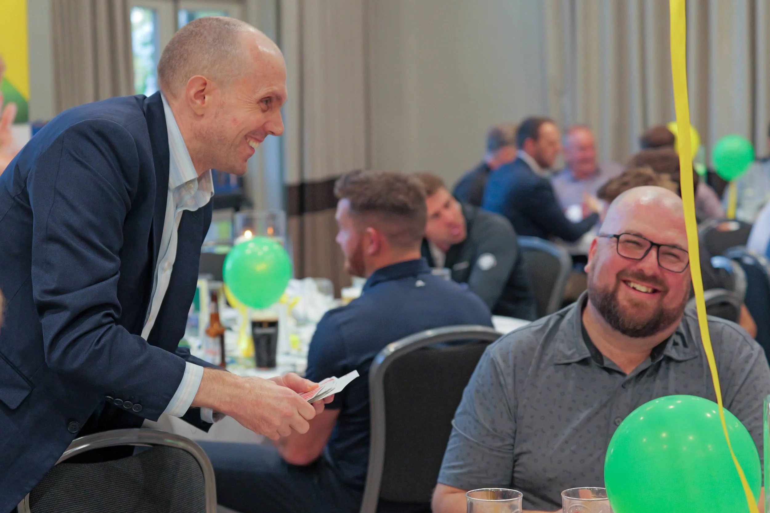 David Richards, wearing a blue suit, is performing a card trick  to a man who is seated at an event, who is laughing and enjoying himself.
.