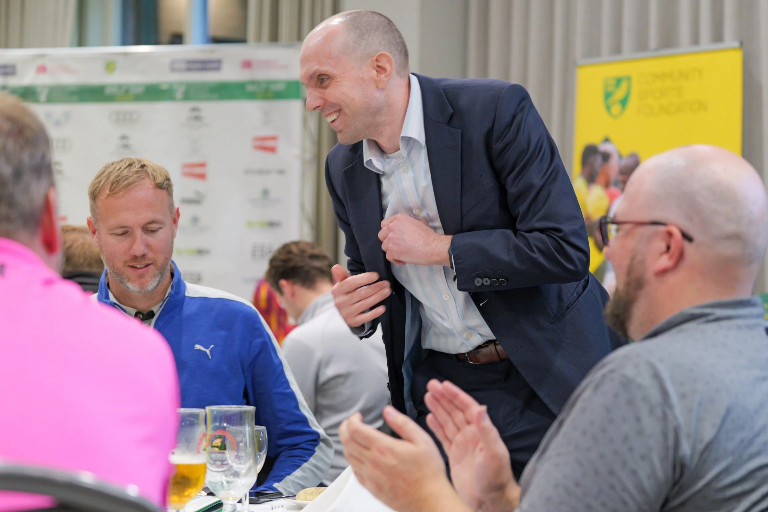 A group of men sitting at a table, with a man in a suit standing and smiling, in front of a yellow and green sign and a backdrop with various logos, at an indoor event.