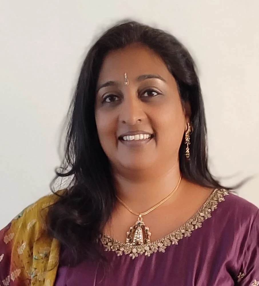 A woman with dark hair wearing traditional Indian attire, a purple saree with gold embroidery, gold jewelry, and a decorative bindi on her forehead, smiling against a plain light-colored background. Internal realisation
